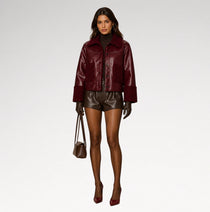 A model stands against a light gray studio background wearing a glossy dark burgundy cropped jacket over a black turtleneck, shiny dark brown leather shorts, black gloves, burgundy heels, and carrying a small brown handbag.
