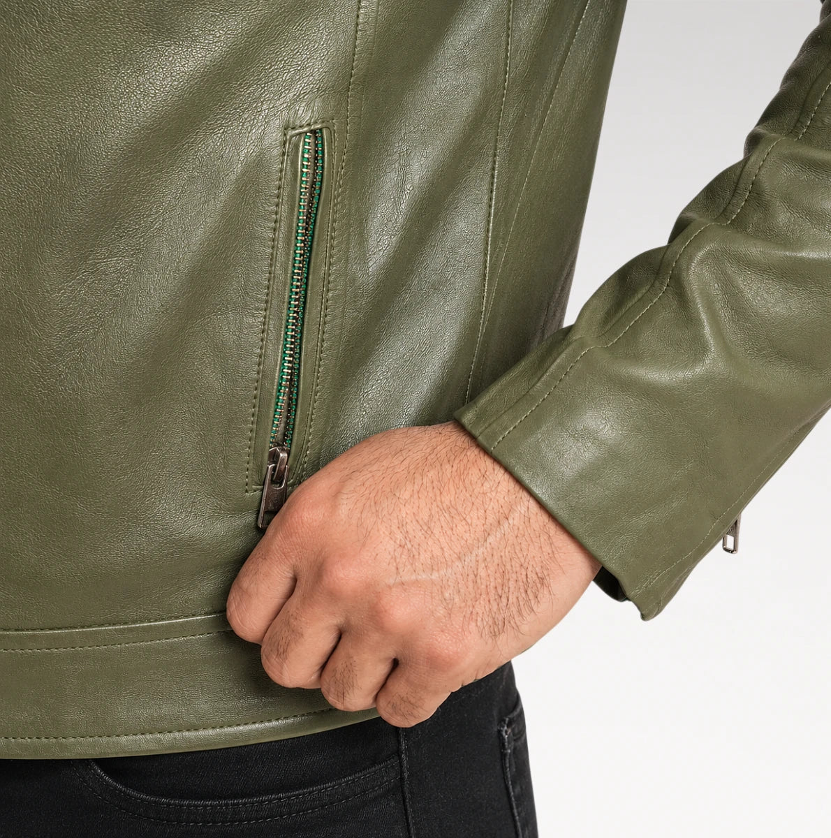 Close-up of a person wearing an olive green faux leather jacket, showing a vertical zippered pocket on the side and a hand resting near the zipper, paired with black jeans against a white background.
