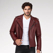 A man wearing a burgundy leather motorcycle jacket with quilted sleeves and diagonal zippers, layered over a white t-shirt and dark jeans, against a light gray studio background.