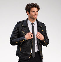 A man wearing a black quilted leather biker jacket with gold-tone hardware, styled with a white dress shirt and black tie, against a light gray studio background.
