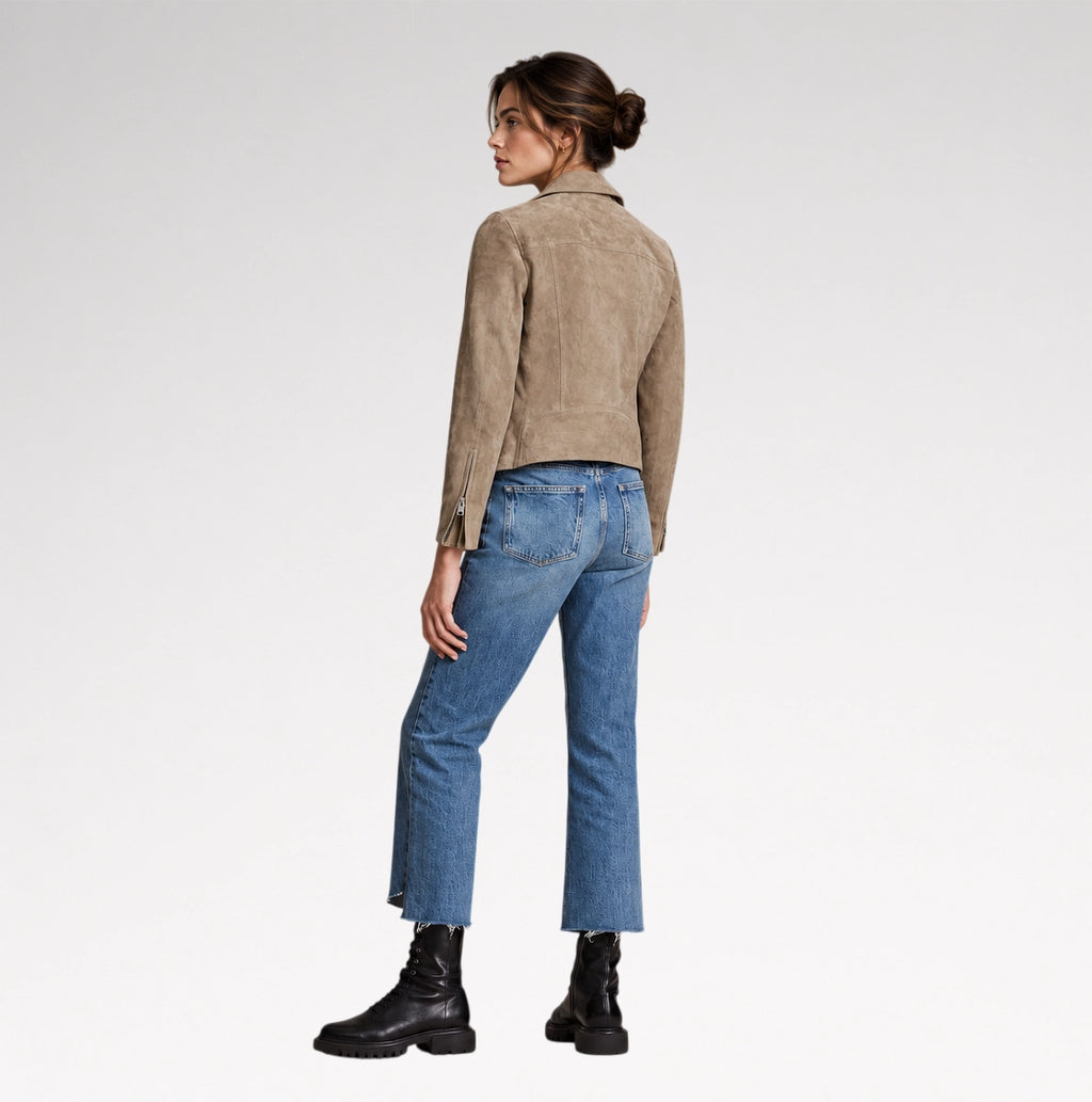 A woman shown from the back wearing a taupe suede cropped jacket, blue jeans, and black boots against a light gray studio background.