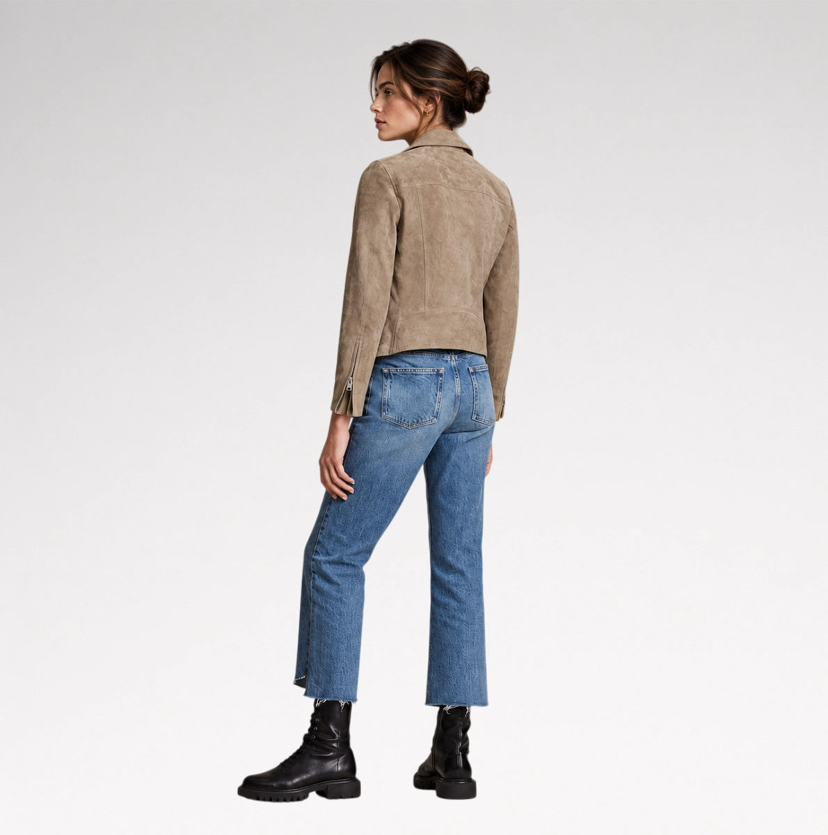 A woman shown from the back wearing a taupe suede cropped jacket, blue jeans, and black boots against a light gray studio background.