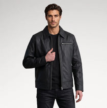 A man wearing a black leather motorcycle jacket with a zippered chest pocket, styled over a black t-shirt and dark jeans, photographed against a light gray studio background.