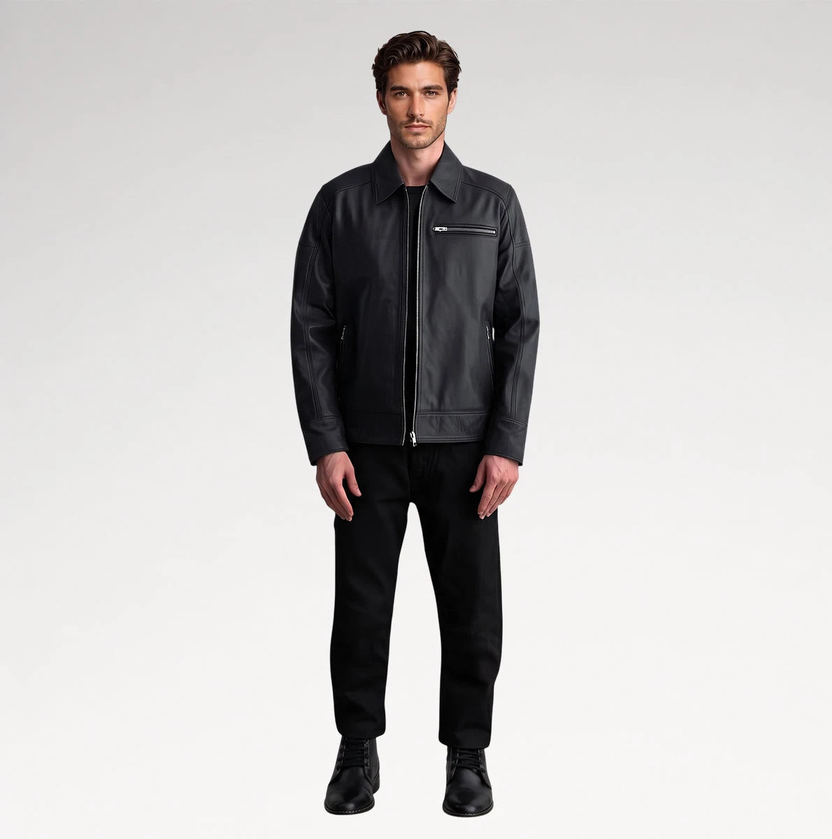 A man modeling a black leather jacket with a zippered chest pocket, paired with black trousers and black shoes, against a light gray studio background.