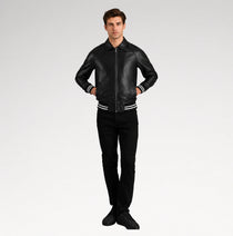 A man modeling a black leather bomber jacket with white stripe details on the cuffs and hem, paired with black pants and black sneakers, against a light gray studio background.