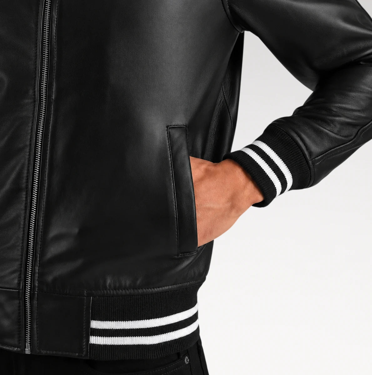 Close-up of a black leather jacket worn by a model, showing the front zipper, side pocket, ribbed knit cuffs with white stripes, and matching striped hemband against a light gray studio background.