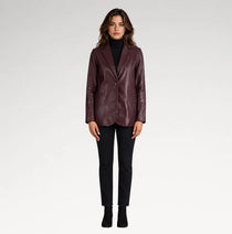 A woman modeling a dark burgundy leather blazer over a black turtleneck, paired with black pants and black ankle boots, against a light gray studio background.