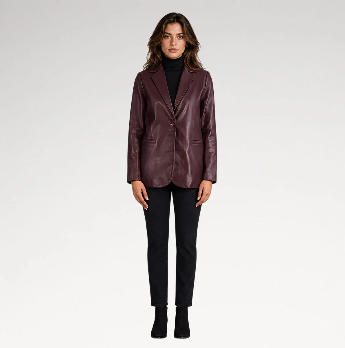 A woman modeling a dark burgundy leather blazer over a black turtleneck, paired with black pants and black ankle boots, against a light gray studio background.