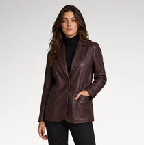 A woman modeling a dark brown faux-leather blazer over a black turtleneck and black pants, photographed against a light gray studio background.