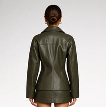 A woman viewed from the back wearing a fitted, textured faux-leather jacket in dark olive green, set against a plain light gray studio background.
