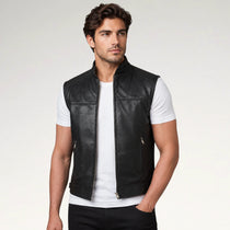 A man wearing a black leather motorcycle vest over a white t-shirt and black pants, photographed against a light gray studio background.