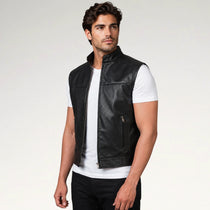 A man wearing a black leather vest over a white t-shirt and black pants, standing against a light gray studio background.
