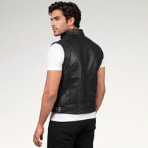 A man wearing a black leather sleeveless vest over a white t-shirt and black pants, shown from a three-quarters back view against a light gray studio background.