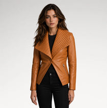 A woman modeling a tan leather moto jacket with a quilted asymmetrical yoke and silver zippers, paired with a black top and black jeans, against a light gray studio background.