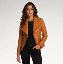 A woman modeling a tan leather moto jacket with a quilted yoke and asymmetrical collar, worn over a black top and black jeans against a light gray studio background.