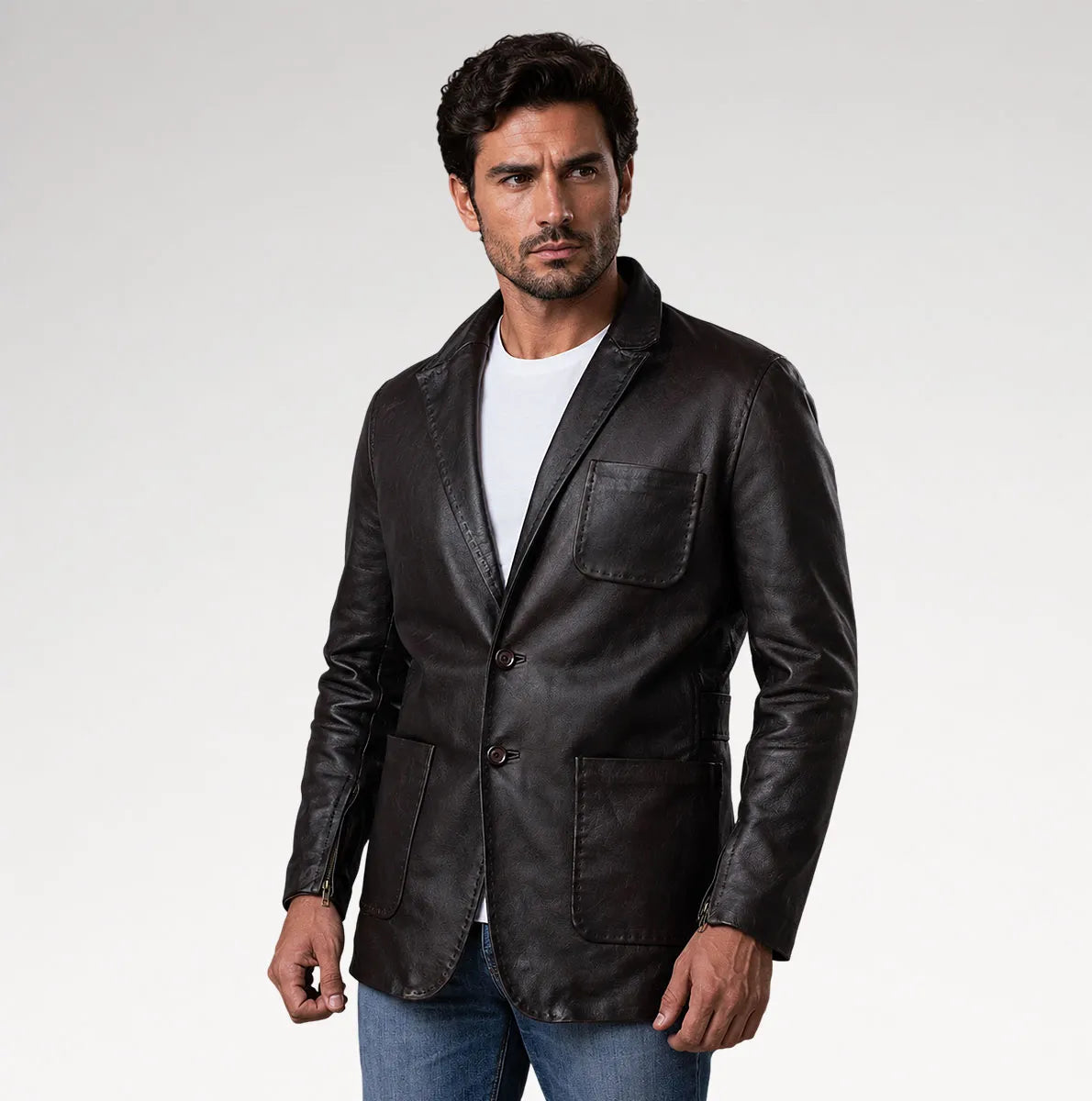 A man wearing a black leather blazer over a white t-shirt and blue jeans, standing against a light gray studio background.