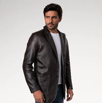 A man wearing a dark brown leather blazer over a white t-shirt and jeans, standing against a light gray studio background.