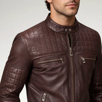 A close-up of a man wearing a dark brown quilted leather jacket with silver zippers, set against a plain light gray studio background.
