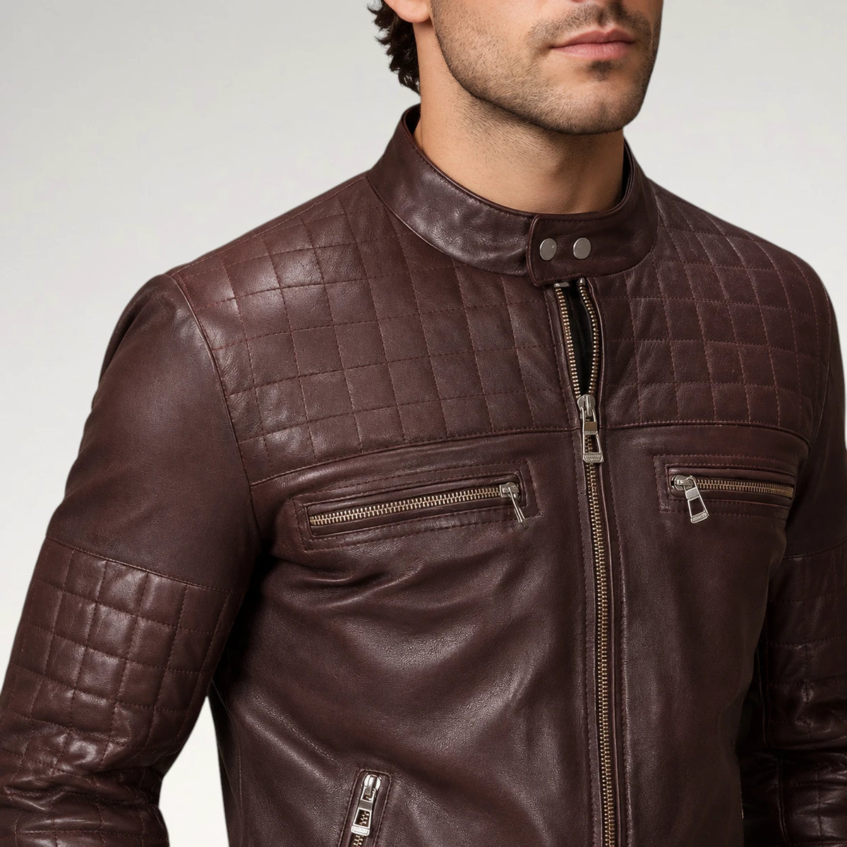 A close-up of a man wearing a dark brown quilted leather jacket with silver zippers, set against a plain light gray studio background.