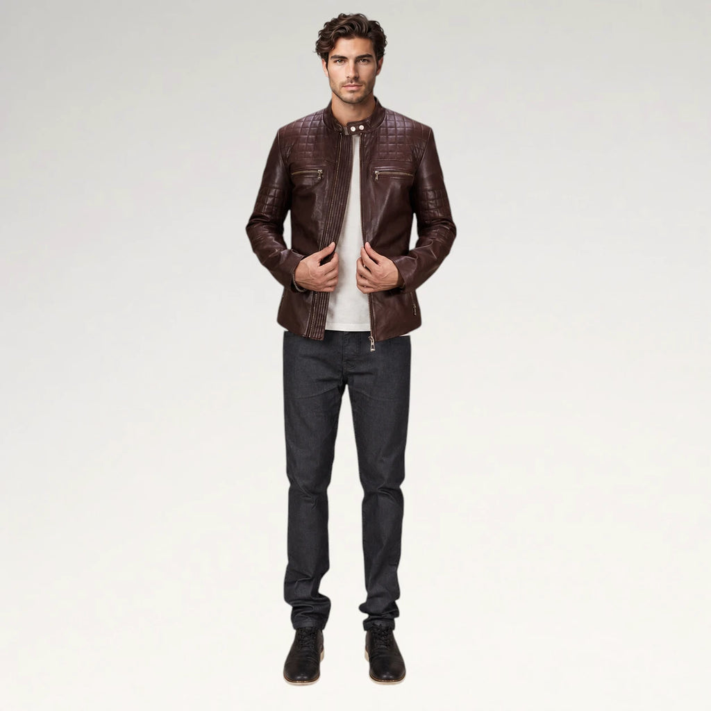 A man modeling a dark brown leather jacket with a subtle embossed pattern, worn over a white t-shirt and dark gray jeans, against a plain light gray studio background.