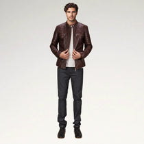 A man modeling a dark brown leather jacket with a subtle embossed pattern, worn over a white t-shirt and dark gray jeans, against a plain light gray studio background.