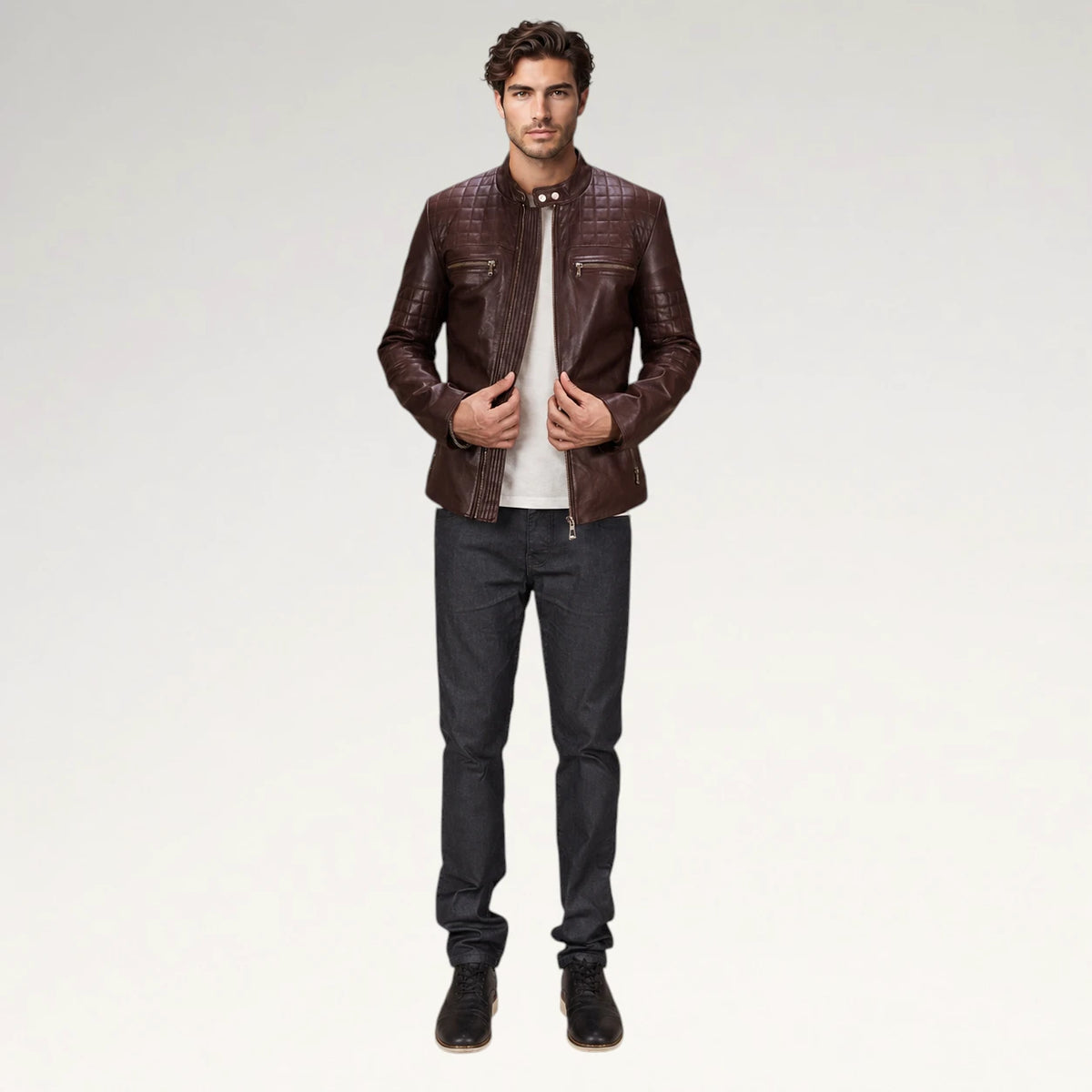 A man modeling a dark brown leather jacket with a subtle embossed pattern, worn over a white t-shirt and dark gray jeans, against a plain light gray studio background.