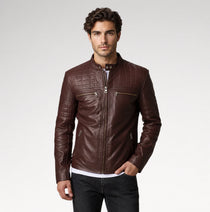 A man wearing a dark brown leather jacket with quilted shoulder panels, a front zipper, and zippered chest pockets, paired with dark jeans against a light gray studio background.