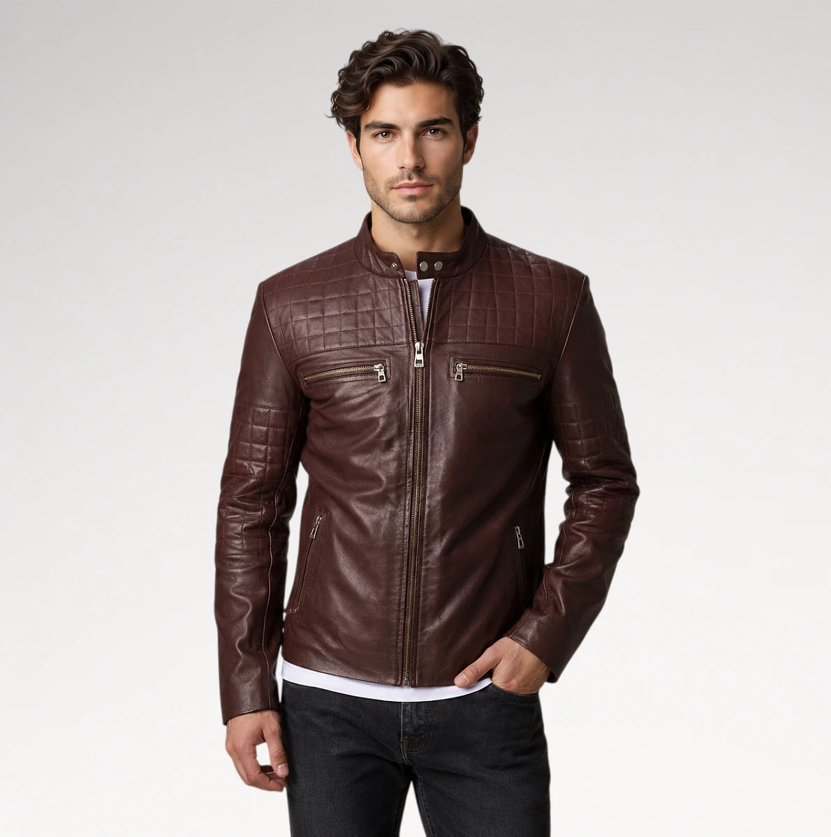 A man wearing a dark brown leather jacket with quilted shoulder panels, a front zipper, and zippered chest pockets, paired with dark jeans against a light gray studio background.