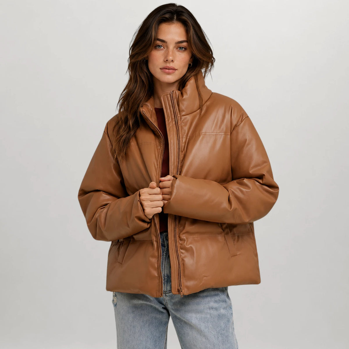 A woman modeling a cropped, quilted faux-leather jacket in a warm camel brown color, styled with light-wash blue jeans over a dark top, against a neutral light gray studio background.