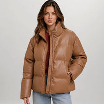 A woman wearing a cropped, quilted faux-leather puffer jacket in a warm camel brown color, styled over a dark red top and light blue jeans, against a neutral light gray background.