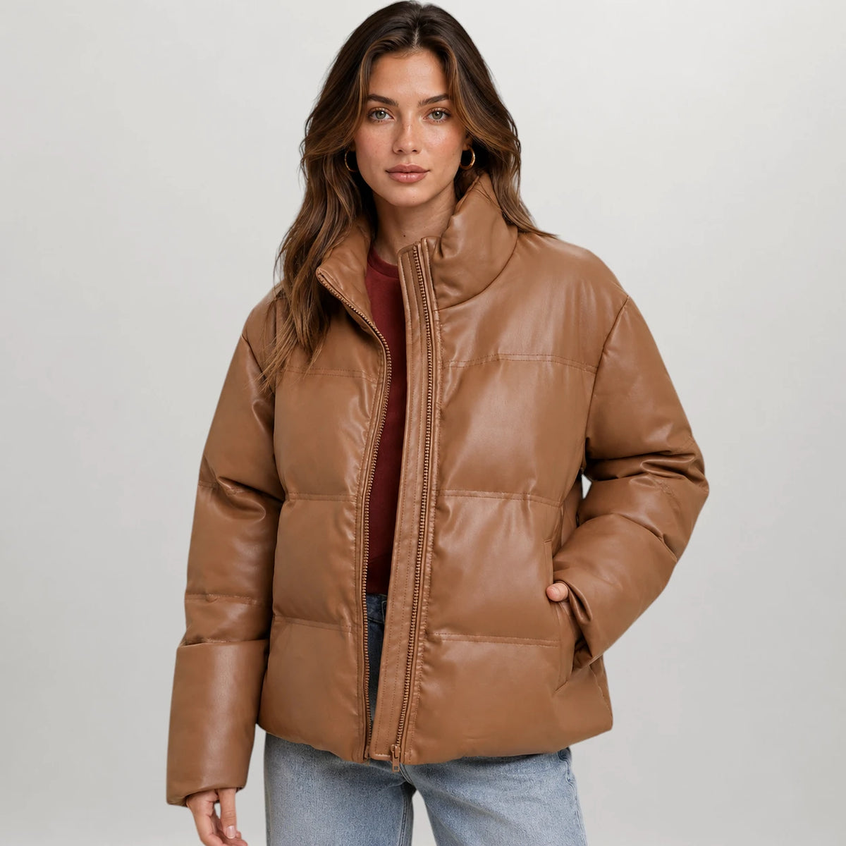 A woman wearing a cropped, quilted faux-leather puffer jacket in a warm camel brown color, styled over a dark red top and light blue jeans, against a neutral light gray background.