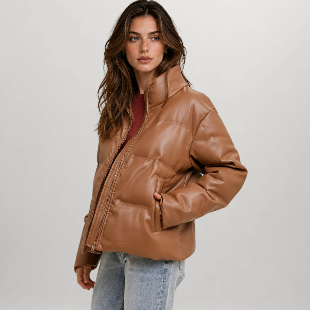 A woman modeling a cropped, shiny brown faux-leather puffer jacket over a dark red top and light blue jeans, photographed against a neutral light gray studio background.