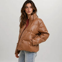 A woman modeling a cropped, shiny brown faux-leather puffer jacket over a dark red top and light blue jeans, photographed against a neutral light gray studio background.