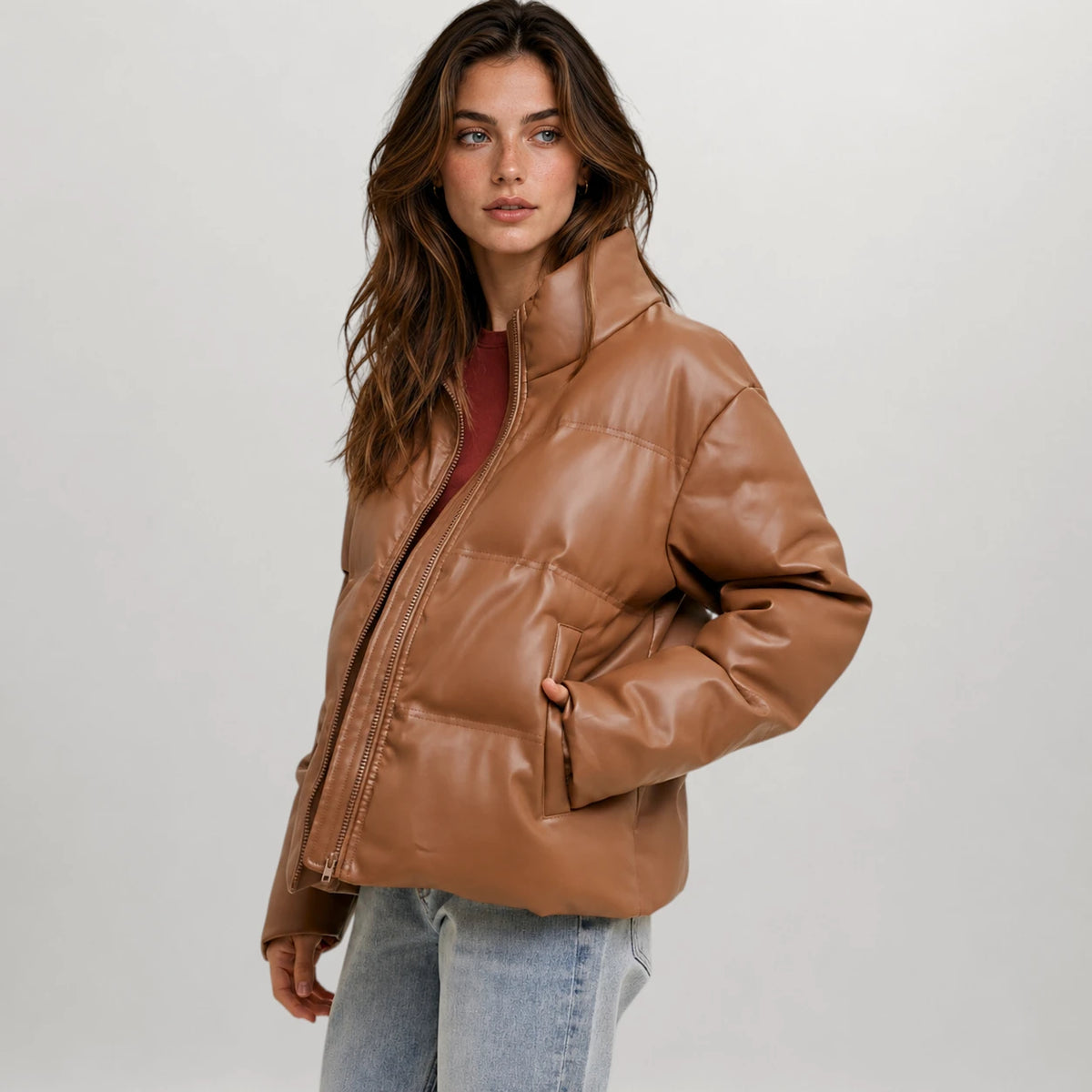 A woman modeling a cropped, shiny brown faux-leather puffer jacket over a dark red top and light blue jeans, photographed against a neutral light gray studio background.