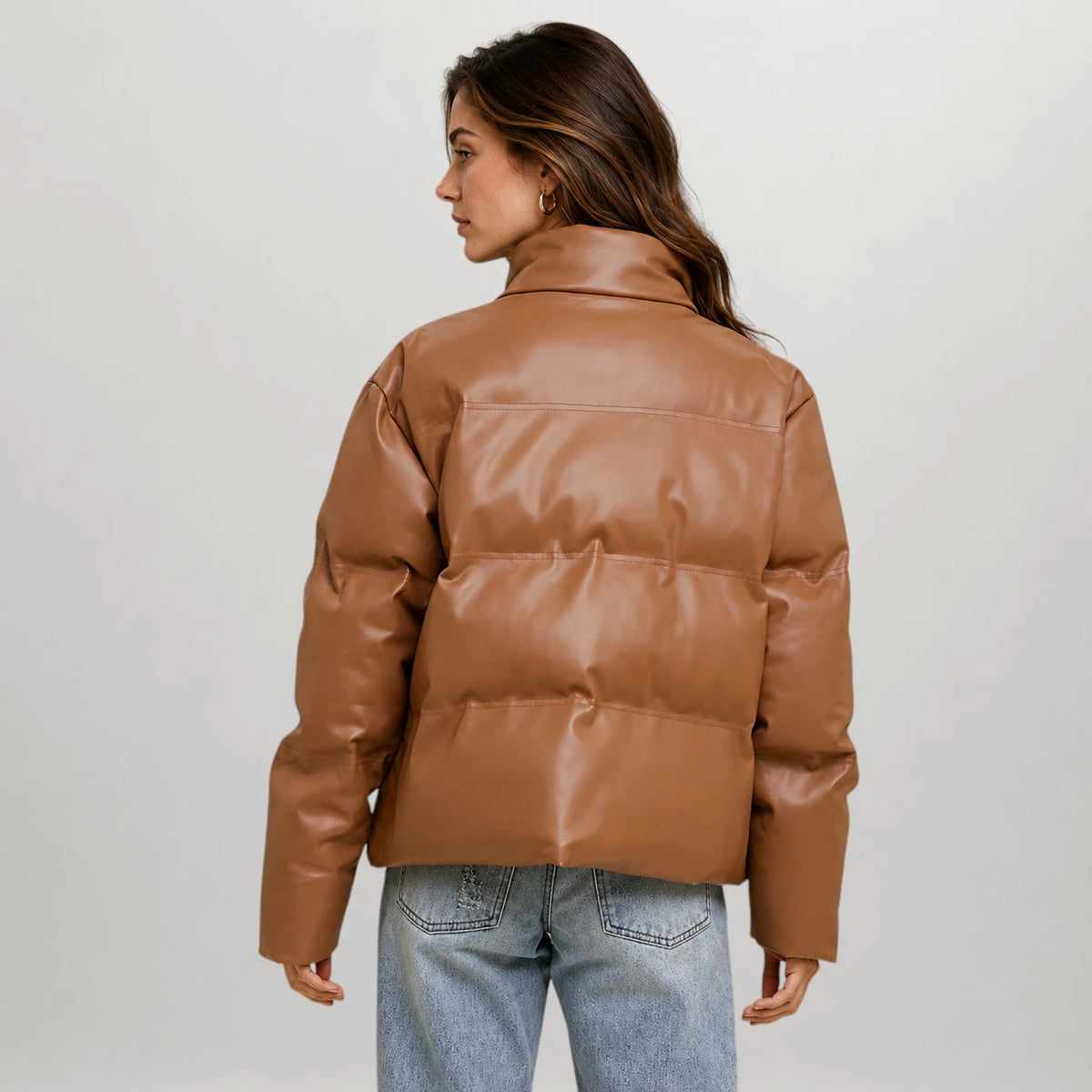 A woman shown from the back wearing a cropped, quilted puffer jacket in smooth tan-brown faux leather, paired with light-wash blue jeans, against a light gray studio background.