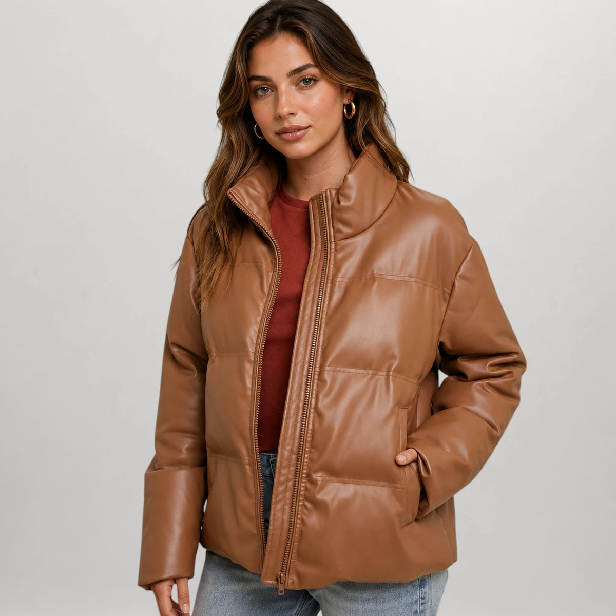 A woman wearing a brown faux-leather puffer jacket over a rust-colored top and blue jeans, photographed against a light gray studio background.