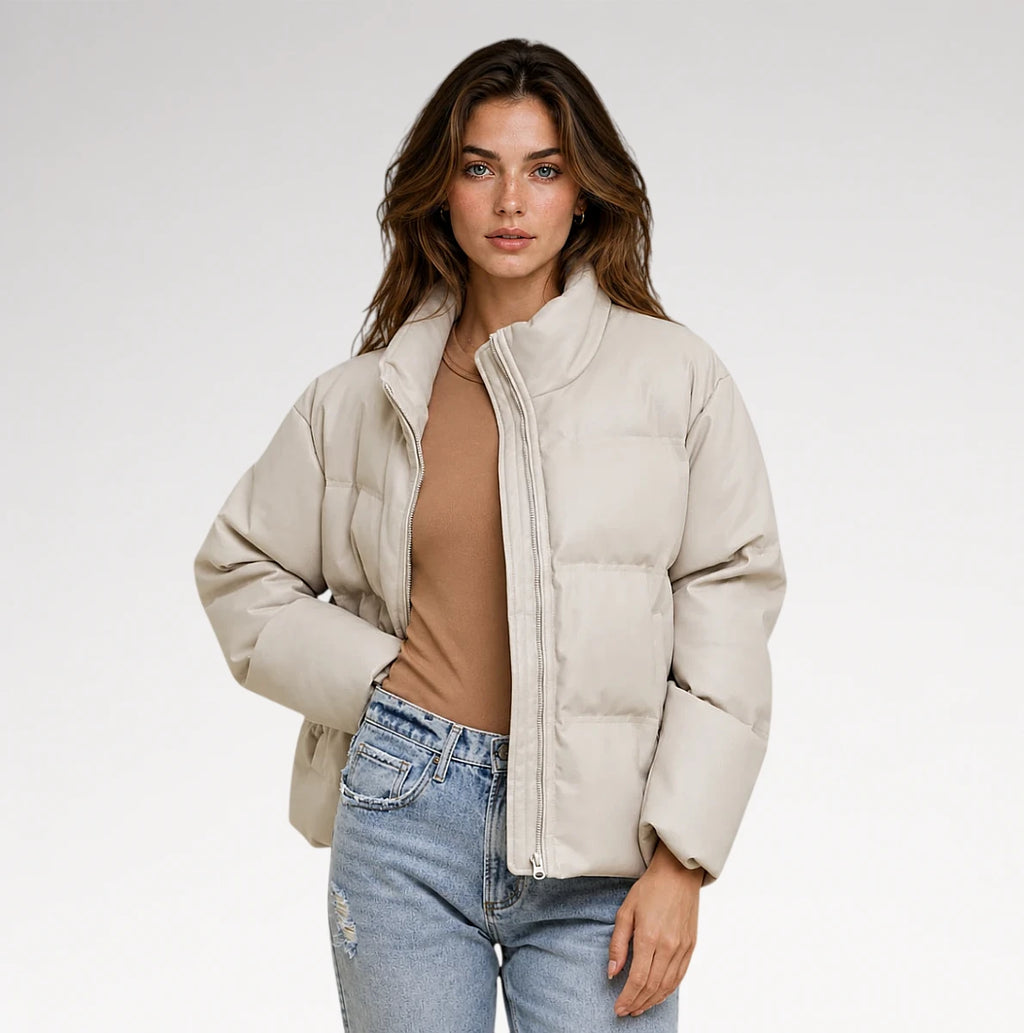 A woman modeling a light beige puffer jacket over a brown top and light blue jeans, photographed against a clean white studio background.
