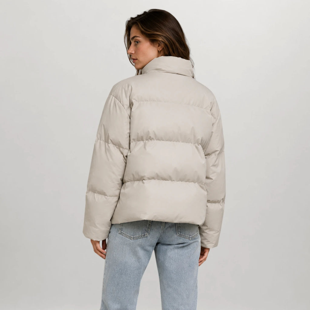 A woman shown from the back wearing a light beige puffer jacket and light blue jeans, photographed against a plain light gray studio background.