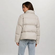 A woman shown from the back wearing a light beige puffer jacket and light blue jeans, photographed against a plain light gray studio background.