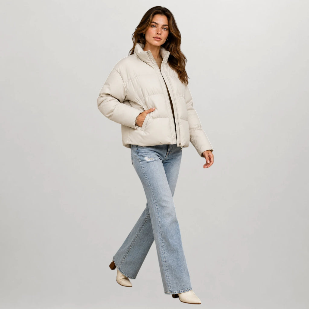 A woman modeling a cropped off-white puffer jacket with a black zipper, paired with light-wash blue jeans and cream ankle boots, against a solid light gray background.