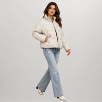 A woman modeling a cropped off-white puffer jacket with a black zipper, paired with light-wash blue jeans and cream ankle boots, against a solid light gray background.