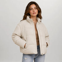 A woman modeling a cream-colored puffer jacket with a high stand-up collar and front zipper, paired with light-wash blue jeans, against a light gray studio background.