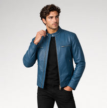 A man wearing a blue leather jacket over a black shirt and black pants, standing against a light gray studio background.