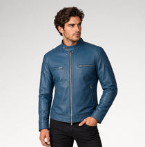 A man wearing a blue leather motorcycle jacket with a stand collar, front zipper, and zippered chest pockets, paired with black pants, photographed against a light gray studio background.