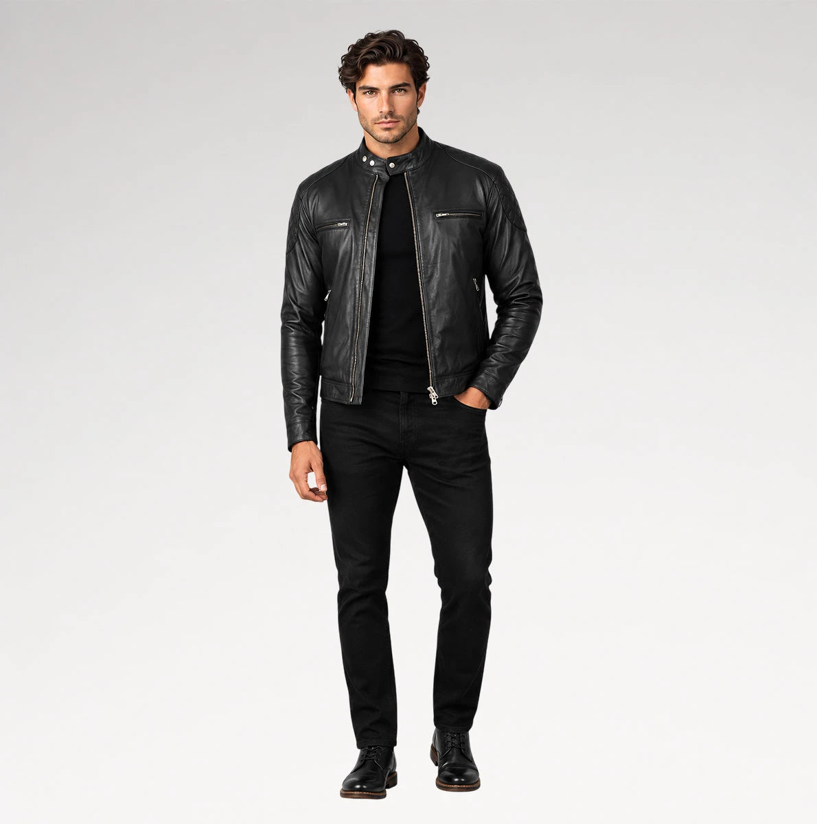 A man modeling a black leather motorcycle jacket over a black shirt and black jeans, standing against a light gray studio background.