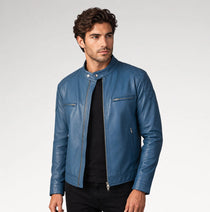 A man wearing a blue leather moto jacket over a black shirt and black pants, standing against a light gray studio background.