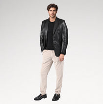 A man modeling a black leather blazer over a black t-shirt, paired with beige trousers and black leather loafers, against a light gray studio background.