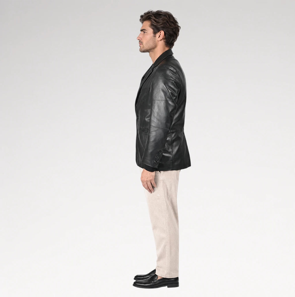 A man in profile wearing a black leather blazer, beige trousers, and black loafers, standing against a light gray studio background.