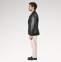 A man in profile wearing a black leather blazer, beige trousers, and black loafers, standing against a light gray studio background.