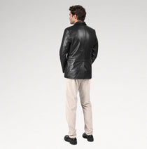 A man wearing a black leather blazer, beige trousers, and black shoes, shown from the back against a light gray studio background.
